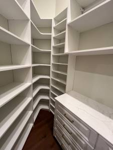 Custom white closet with floor-to-ceiling corner shelving, wraparound open shelves, and quartz drawer unit