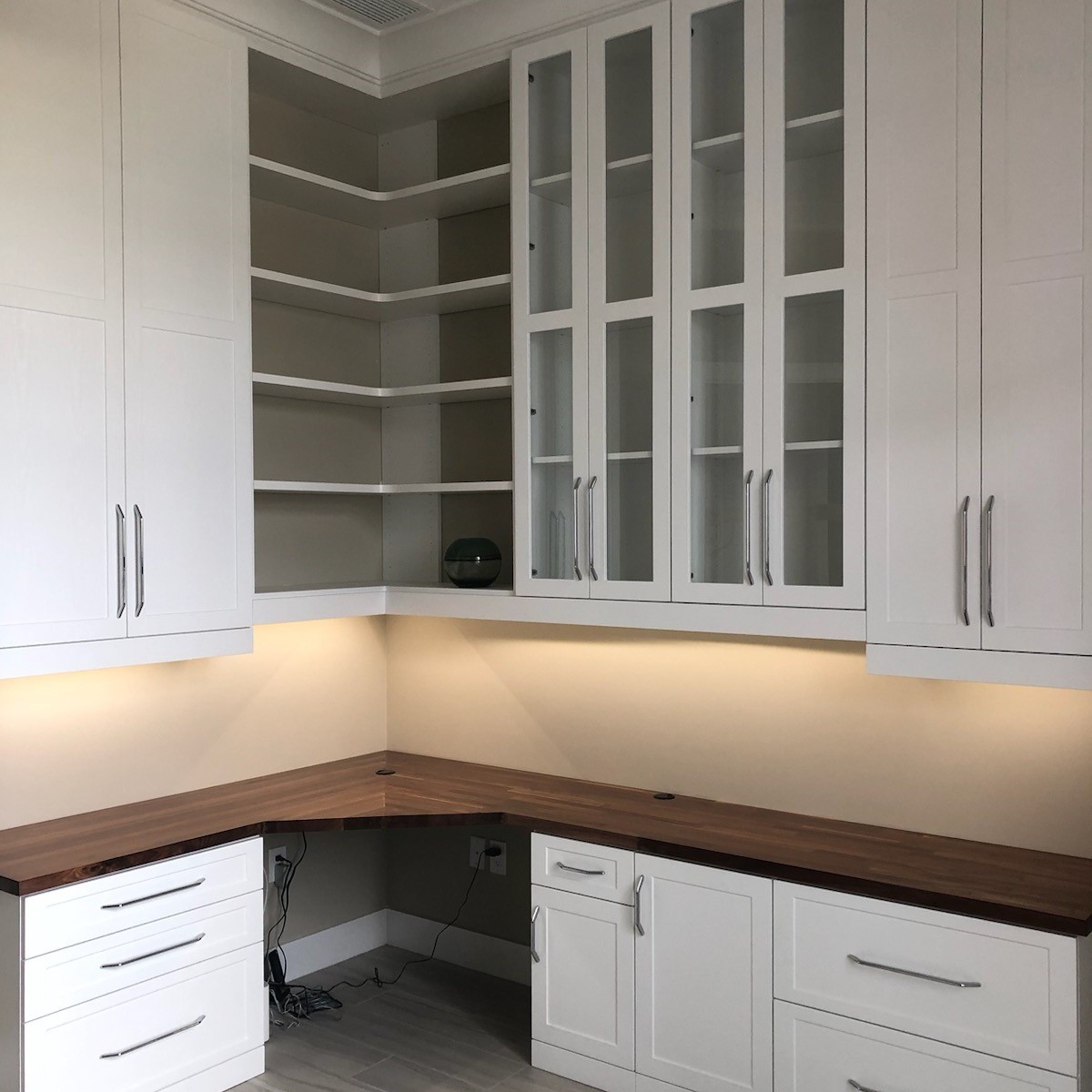 Custom L-shaped home office with white shaker cabinetry, walnut butcher block countertop, glass-front upper cabinets, corner open shelving, and under-cabinet lighting