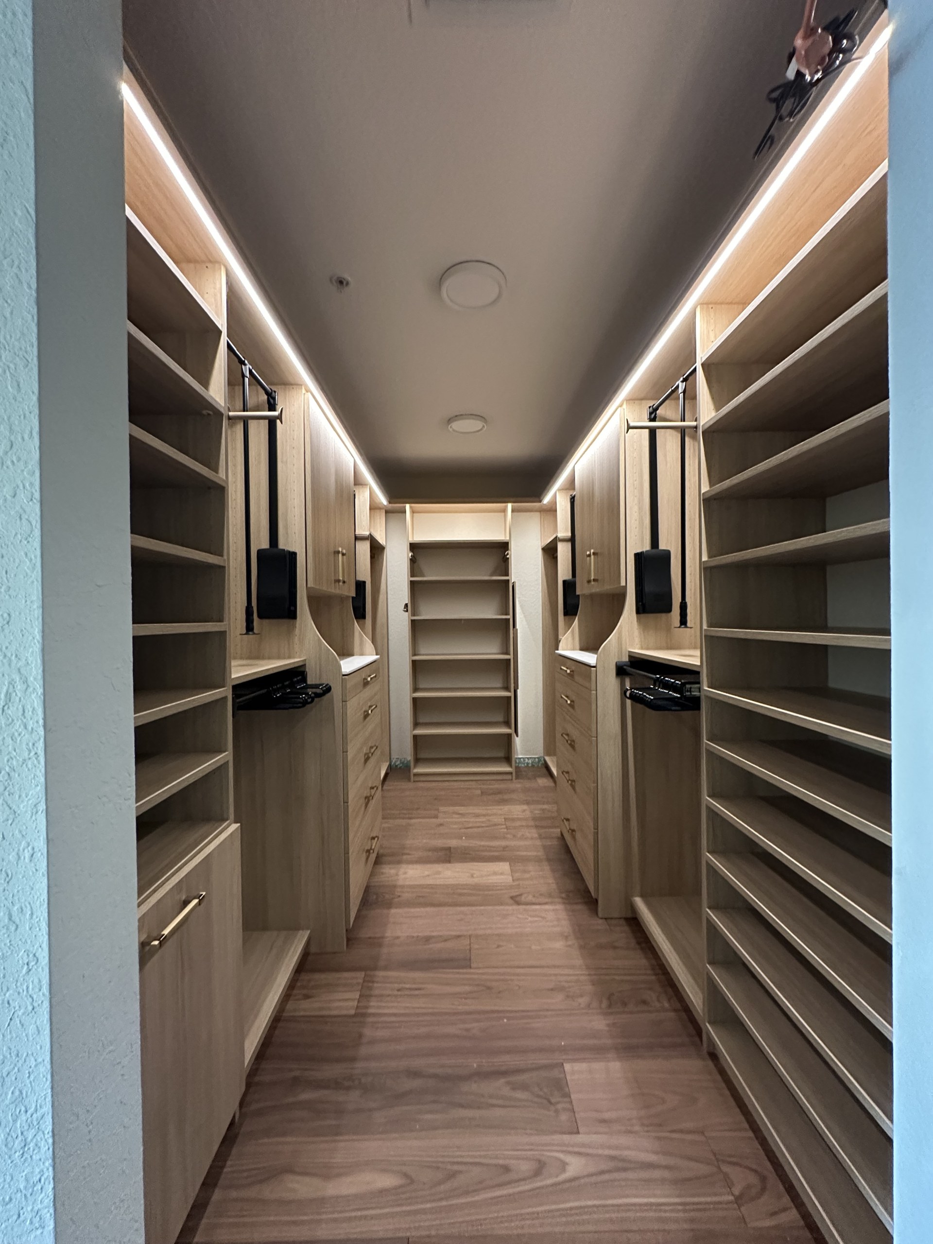 Luxury aisle-style walk-in closet in light oak with pull-down wardrobe bars, LED strip ceiling lighting, and built-in valet accessories, More Space Place Florida