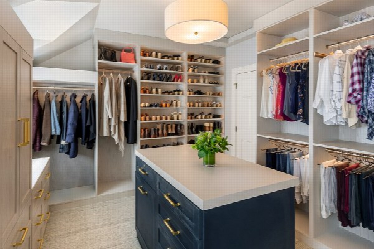 Custom walk-in closet with two-tone configuration featuring light linen woodgrain perimeter cabinetry and contrasting navy shaker island with brass pulls, dedicated shoe shelving tower, open hanging sections, and light countertop surface